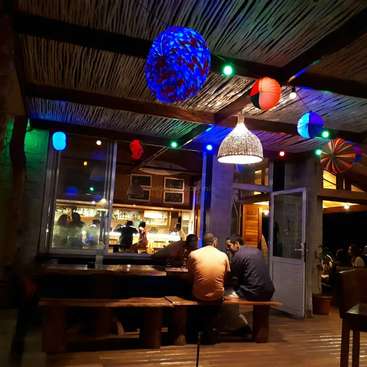 A imagem mostra um restaurante aconchegante com piso e teto de madeira, com uma grande janela, lanternas coloridas e clientes sentados à mesa, criando um ambiente acolhedor.