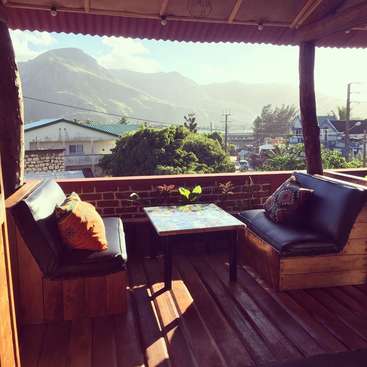 A imagem mostra uma varanda aconchegante com piso e teto de madeira, com uma pequena mesa e duas cadeiras, com vista para uma paisagem montanhosa serena e uma cidade pitoresca abaixo.