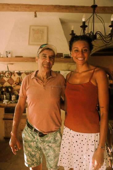 Un homme âgé portant une casquette et un short se tient à côté d'une jeune femme souriante vêtue d'un haut rouge et d'une jupe à pois, à l'intérieur d'une cuisine confortable.