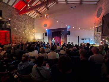A lively audience fills a cozy, rustic venue with exposed stone walls, warmly lit by string lights. A performer stands on stage, engaging the crowd.
