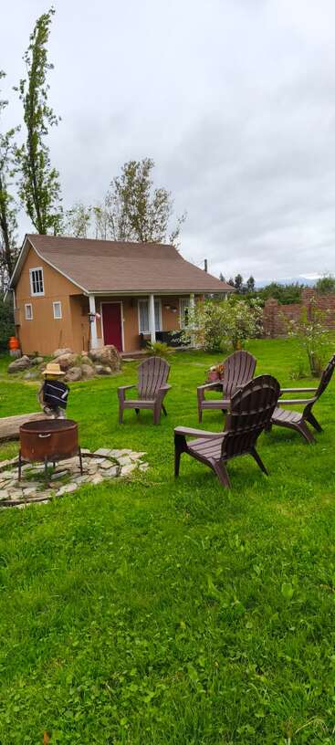 Una acogedora casita de campo con porche se encuentra en un exuberante jardín. Las sillas Adirondack rodean una hoguera que invita a relajarse en plena naturaleza.