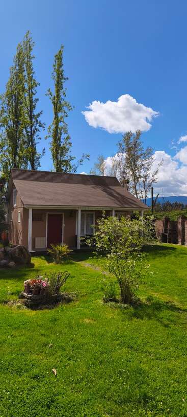 Una acogedora casa con tejado marrón descansa apaciblemente bajo un cielo azul. La exuberante hierba verde, los árboles, las flores y las esponjosas nubes blancas completan la escena.