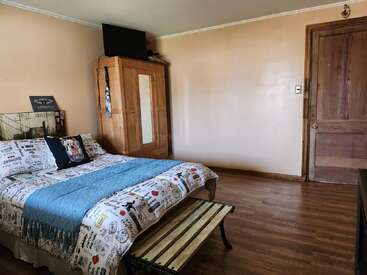 Acogedora habitación con suelo de madera, cama de matrimonio con ropa de cama estampada, armario de madera, televisión encima, un banco a los pies y una puerta de madera.