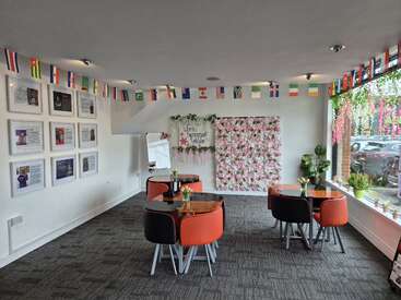 Cette photo montre une pièce lumineuse et moderne, avec des petites tables, des compositions florales, des drapeaux du monde entier, des portraits accrochés au mur et de grandes fenêtres ornées de plantes en pot. L'ambiance y est chaleureuse.