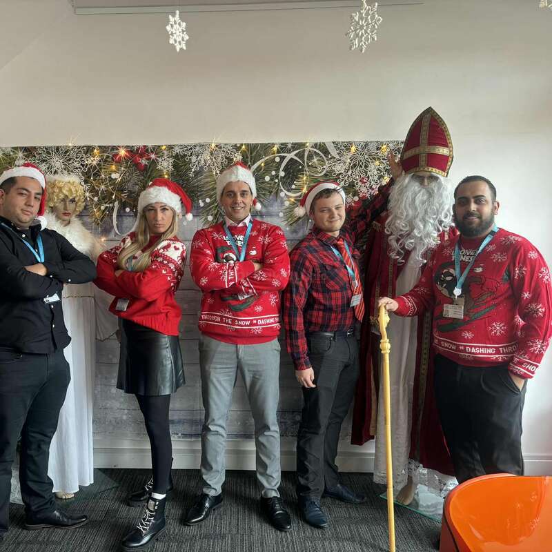 Six personnes posent en tenue de fête, avec notamment des bonnets de Père Noël et des pulls de Noël, aux côtés d'une figurine de Saint Nicolas. Des flocons de neige et des décorations de Noël sont suspendus en arrière-plan.