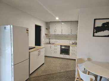 Cette cuisine moderne est équipée de meubles blancs, d'un réfrigérateur, d'un four et d'appareils électroménagers de comptoir. Elle comprend une petite table à manger avec des chaises, une décoration minimaliste et un sol aux tons neutres.