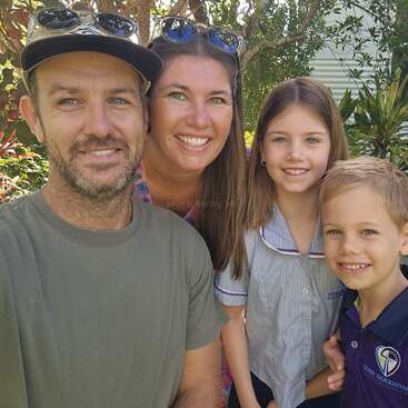 Une joyeuse famille de quatre personnes pose en plein air, arborant un sourire chaleureux. Les parents se tiennent derrière leurs deux enfants, entourés de verdure et baignés de soleil, profitant ensemble d'une belle journée.