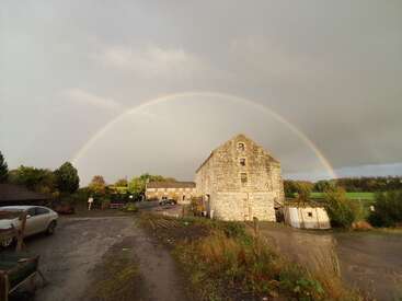Un magnifique arc-en-ciel traverse un ciel nuageux au-dessus d'une vieille bâtisse en pierre, de champs verdoyants et d'un chemin boueux, capturant une scène rurale paisible.