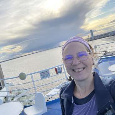A smiling person with glasses and a headband is on a boat deck. The background shows water, distant buildings, and a dramatic cloudy sky at sunset.