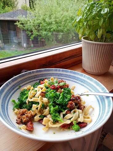 A plate of pasta with broccoli, sausage, and sun-dried tomatoes sits by a window. A basil plant in a ceramic pot is nearby, overlooking a green garden.