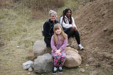 Three people are sitting on large rocks outdoors. The background is grassy and earthy. They appear relaxed and happy, enjoying a casual moment together in nature.