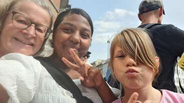 Three people are smiling and posing for a selfie outdoors on a sunny day, making peace signs. A fourth person stands with their back to the camera.