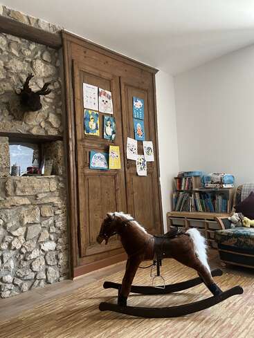 Una acogedora habitación cuenta con una pared rústica de piedra, un armario de madera decorado con dibujos infantiles, estanterías, una silla acolchada y un caballito de balancín marrón sobre una alfombra de textura.
