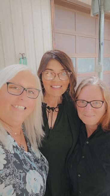 Trois femmes se tiennent côte à côte et sourient largement à l'appareil photo. Elles portent toutes des lunettes et des vêtements décontractés ; elles semblent heureuses et apprécient la compagnie les unes des autres, peut-être lors d'une réunion en plein air.