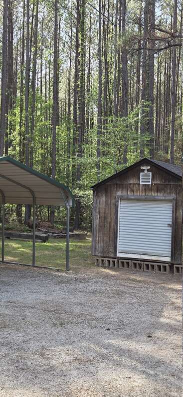 Une cabane rustique en bois, dotée d'une porte roulante en métal, se trouve à côté d'un abri de voiture en métal, entourée de grands pins et d'une clairière de gravier dans les bois.