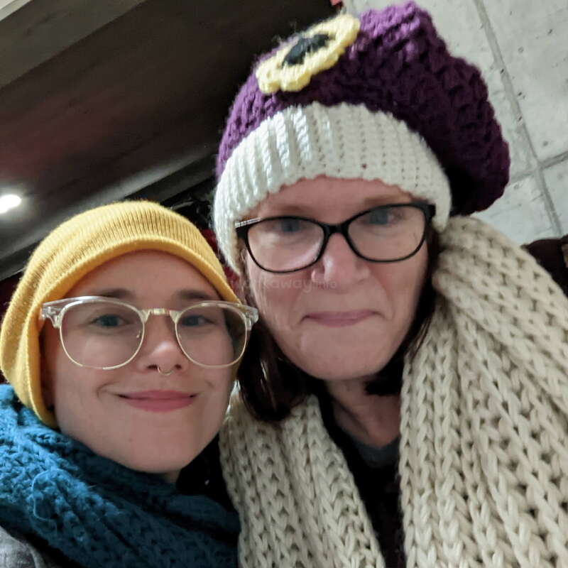 Deux personnes bien emmitouflées sourient pour un selfie. Toutes deux portent des lunettes, des bonnets en tricot et des écharpes, créant ainsi une ambiance hivernale chaleureuse, conviviale et joyeuse à l'intérieur.