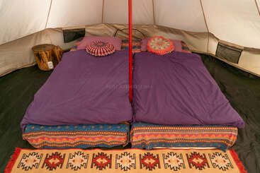 Esta imagem mostra duas camas com roupa de cama roxa e travesseiros coloridos e estampados dentro de uma tenda. Há um poste central, um toco de madeira e um tapete estampado.