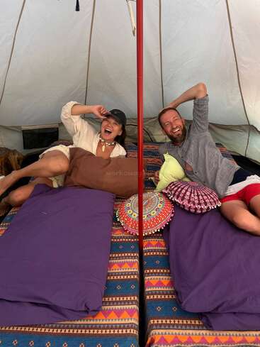 Um homem e uma mulher alegres estão descansando em uma tenda aconchegante. Eles estão rindo, cercados por almofadas e roupas de cama coloridas, desfrutando de um momento relaxante e alegre juntos.