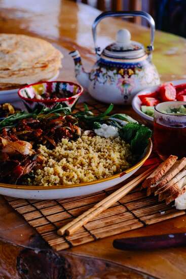 Uma mesa lindamente posta apresenta arroz, legumes salteados, ervas, pão achatado, pão fatiado, salada, chá em um copo, um bule colorido e condimentos.