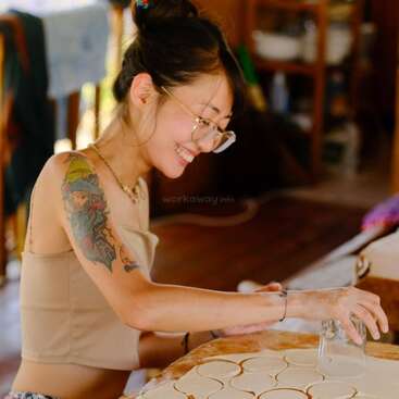 Uma mulher sorridente com óculos e uma tatuagem colorida no braço está usando um copo para cortar círculos de massa em uma mesa de madeira em uma cozinha aconchegante.