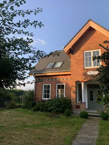 A charming brick house with large windows and a green door stands amidst a lush garden, bathed in warm evening sunlight under a clear blue sky.