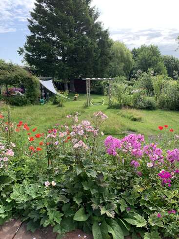 A vibrant garden scene with colorful flowers in the foreground, lush green grass, a swing, a pergola, and tall trees under a partly cloudy sky.