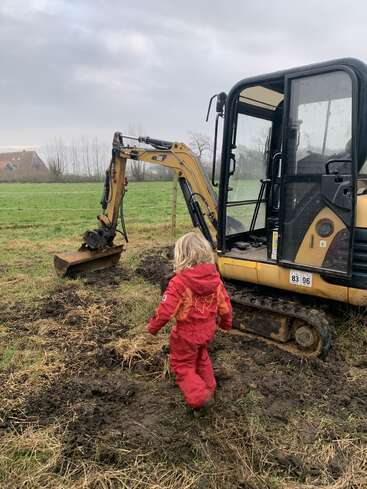 A child in a red rain suit walks toward a yellow excavator on a muddy field, with overcast skies and a distant house in the background.