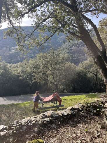 A woman lays a red cloth on a picnic table under large trees, surrounded by green hills and stone walls, enjoying a peaceful, sunny afternoon outdoors.