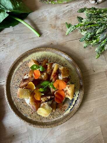 A rustic ceramic plate holds roasted root vegetables garnished with edible orange flowers and fresh greens. Asparagus and leafy greens are arranged on the wooden table nearby.