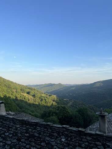This image shows a serene mountain landscape with lush green hills, a stone rooftop in the foreground, and a clear blue sky above, peaceful and calm.