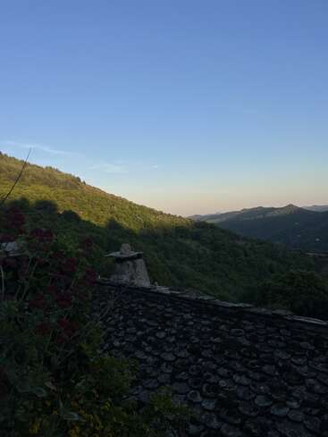 A serene mountain landscape at sunset, green hills stretching into the distance, a rustic tiled roof and chimney in the foreground, framed by dusky blue sky.