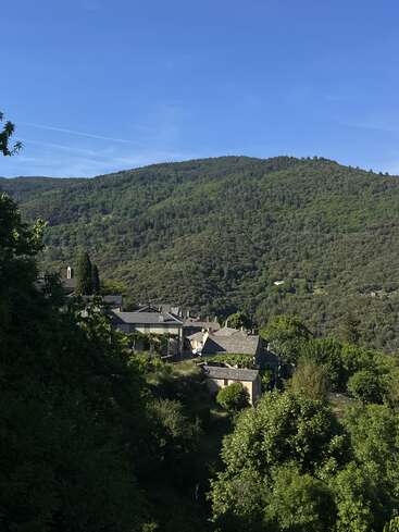 A peaceful village nestles among lush, green hills under a clear blue sky. Stone-roofed houses are surrounded by dense trees and natural beauty.
