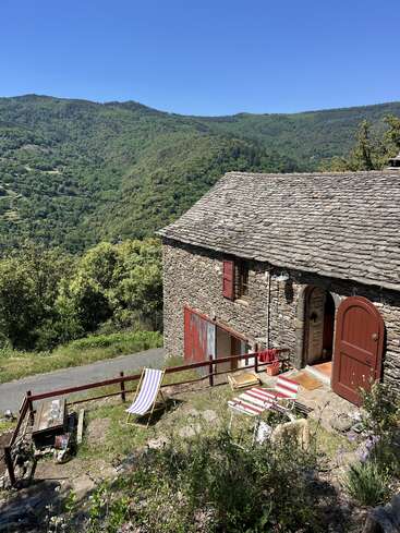 A rustic stone house with red doors sits on a hillside, overlooking lush green mountains. Striped deck chairs and a table invite relaxation under sunny skies.