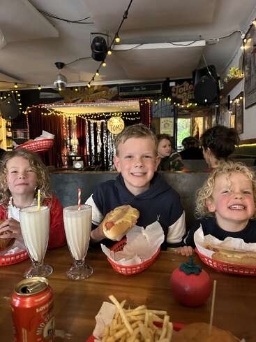 Trois enfants heureux sont assis dans un restaurant animé, souriant devant des milkshakes, des hot-dogs et des frites. Les lumières chaudes et les décorations rétro créent une atmosphère amusante et joyeuse.