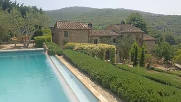 Une villa rustique en pierre se trouve au milieu d'une végétation luxuriante et de collines ondulantes. Au premier plan, une piscine sereine surplombe des haies manucurées, avec une personne assise au bord de la piscine.