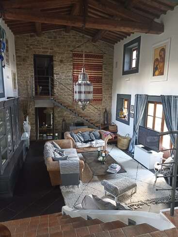 Ce salon confortable présente des murs en pierre, des poutres en bois, un décor éclectique, des canapés confortables, un tapis à motifs, de grandes fenêtres, des textiles vibrants, une télévision, des livres et un éclairage d'ambiance.