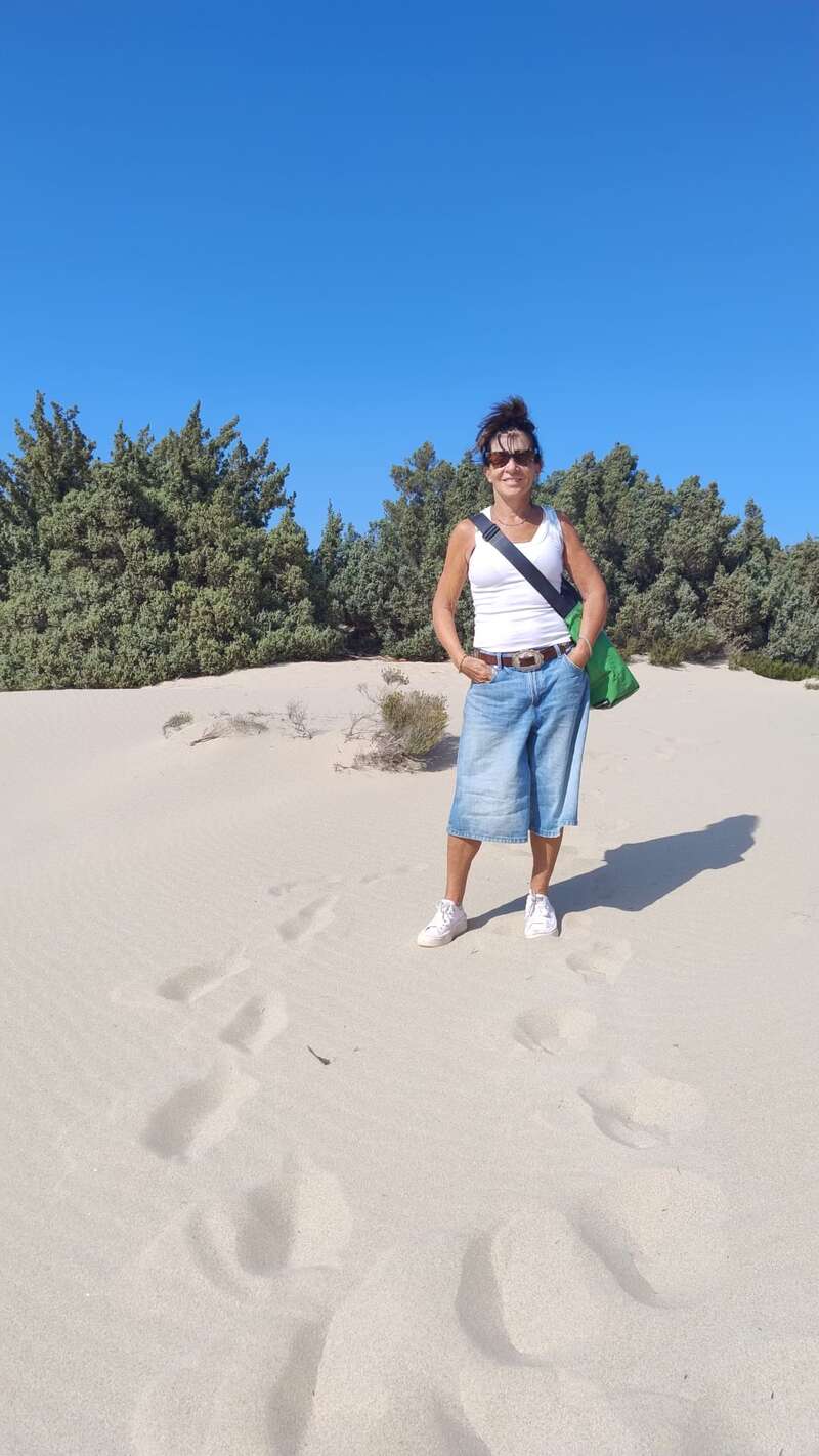 Une femme se tient avec assurance sur des dunes de sable sous un ciel bleu clair, portant des lunettes de soleil, un débardeur blanc, un short en jean et des baskets, avec des arbres verts en arrière-plan.