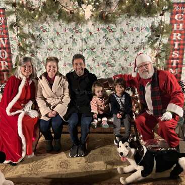 La imagen muestra a una familia de cuatro miembros posando con Papá Noel y la Sra. Noel en un ambiente festivo, rodeados de adornos navideños y un perro de peluche.
