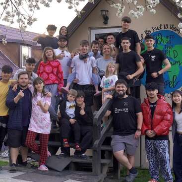 Un groupe hétéroclite de personnes, composé d'enfants et d'adultes, pose joyeusement devant un petit bâtiment, sous des branches d'arbres en fleurs, en souriant et en échangeant des gestes amicaux.