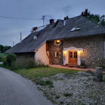 A rustic stone house sits beside a quiet road at dusk. Warm lights glow by the entrance, surrounded by greenery and a peaceful rural landscape.