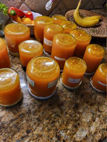 Several glass jars filled with orange marmalade are cooling upside down on a kitchen counter. Nearby, bananas and peppers rest in baskets and bowls.