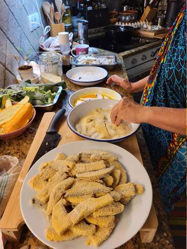 A person is breading sliced vegetables in a cozy kitchen, preparing them for frying. Fresh ingredients and kitchen utensils are neatly arranged on the countertop.