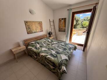 A cozy bedroom with leafy bedding, wooden furniture, and artwork on white walls. Large glass door opens to a sunny terrace with outdoor seating and trees.