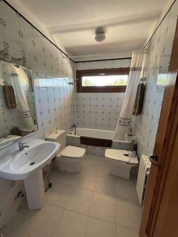 This is a bright bathroom with white patterned tiles, a bathtub with a curtain, a window, sink, mirror, toilet, bidet, towels, and radiator.
