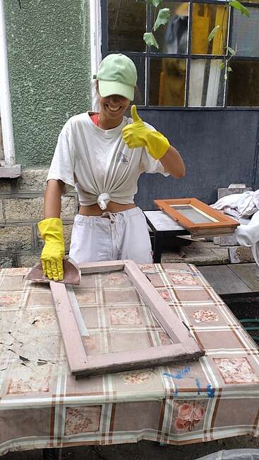 A smiling person wearing a green cap, white clothes, and yellow gloves gives a thumbs-up while sanding a wooden frame outdoors on a patterned tablecloth.