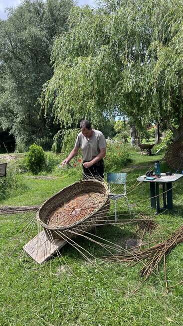 Ein Mann flechtet einen großen Korb im Freien in einem üppigen, grünen Garten, umgeben von Bäumen, einem Tisch, Stühlen und einer Schubkarre im Hintergrund.