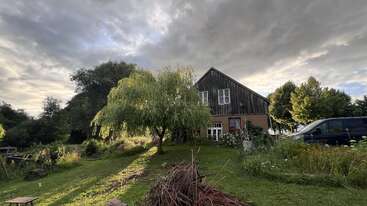 Ein rustikales Haus steht unter einem dramatisch bewölkten Himmel, umgeben von üppigem Grün, Bäumen, Wildblumen, einem Lieferwagen und einem ordentlich gestapelten Stapel Holzstöcke.