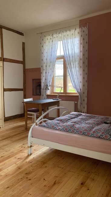 Ein gemütliches, rustikales Schlafzimmer mit Holzböden, einem weißen Metallbett, geblümten Vorhängen, einem hölzernen Schreibtisch und Stuhl am Fenster und natürlichem Sonnenlicht, das hereinströmt.