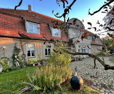 Una encantadora casa antigua con tejado de tejas rojas, rodeada de exuberante vegetación, patio empedrado y plantas en flor, bajo un cielo azul despejado al atardecer.