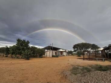 Esta imagen muestra una granja rural bajo un cielo nublado con un arco iris doble muy vivo, árboles, espacios abiertos y un pequeño animal cerca de los edificios.